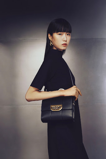 Asian woman with black hair wearing a black dress and gold earrings holding a black leather purse in front of a metallic backdrop.
