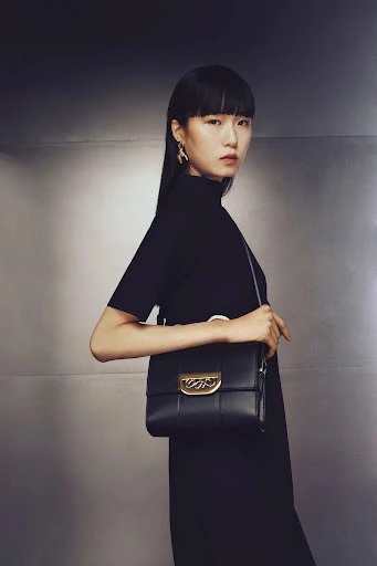 Asian woman with black hair wearing a black dress and gold earrings holding a black leather purse in front of a metallic backdrop.