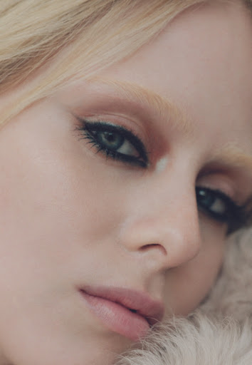 Close-up portrait of a woman with blonde hair and dramatic eye makeup, resting against a soft, furry surface in soft, diffused lighting.