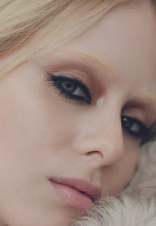Close-up portrait of a woman with blonde hair and dramatic eye makeup, resting against a soft, furry surface in soft, diffused lighting.