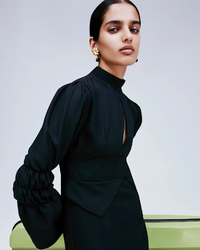 Elegant adult female fashion model poses indoors wearing a sophisticated black dress, accessorized with statement gold earrings.