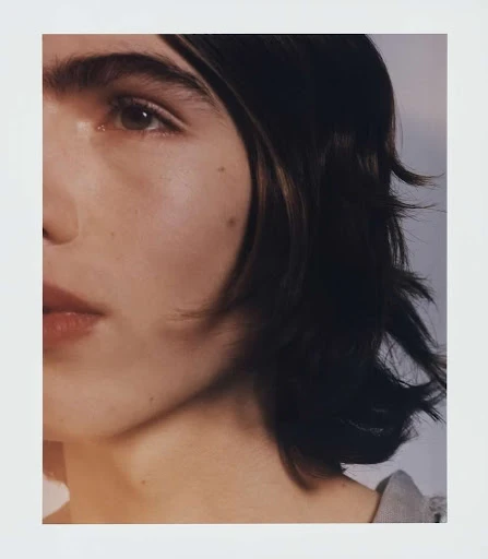 Close-up portrait of a young adult woman with dark hair, neutral expression, and fair skin in a softly lit indoor setting.