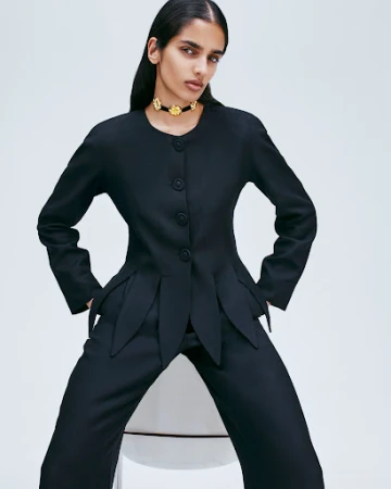 Model wearing a black tailored jacket with petal hem details and a gold floral choker against a seamless white background.