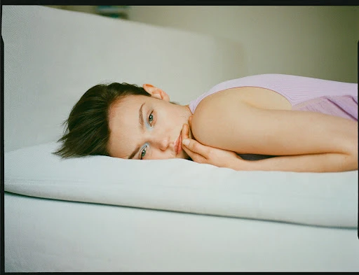 Young woman with short hair rests on a white surface with her hands near her face, wearing a lilac top and light blue eyeshadow.