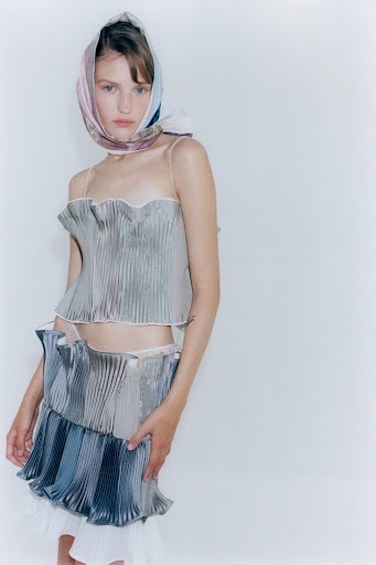 A model poses in a silver, pleated two-piece outfit and floral headscarf against a bright backdrop in a studio fashion shot.