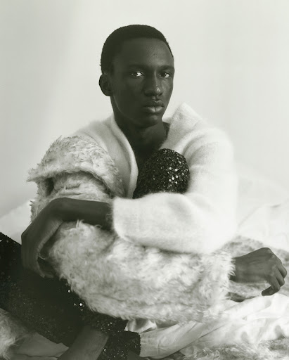 Portrait of a Black male model in a white sweater, seated on a bed with white sheets, holding a black, beaded purse.