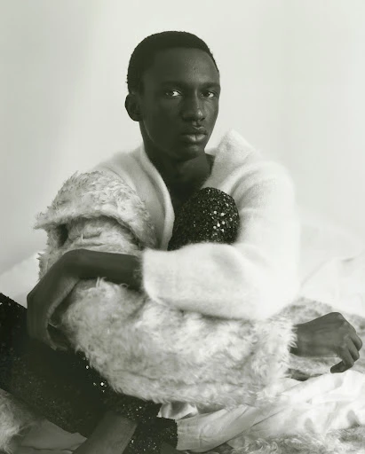 Portrait of a Black male model in a white sweater, seated on a bed with white sheets, holding a black, beaded purse.