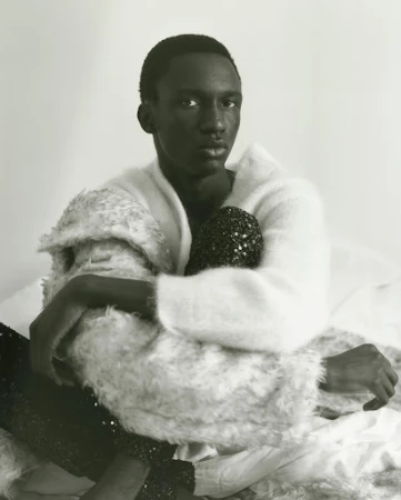 Portrait of a Black male model in a white sweater, seated on a bed with white sheets, holding a black, beaded purse.