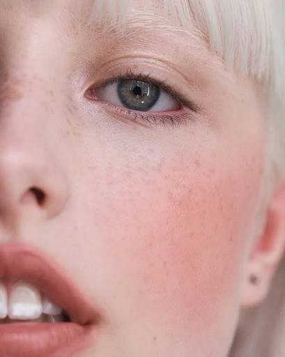 Close-up portrait of a fair-skinned young woman with pale blonde hair, featuring a partial view of her face, a green eye, and parted lips.