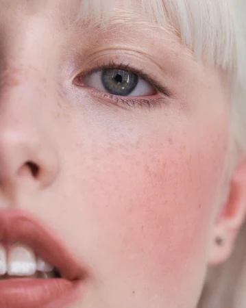 Close-up portrait of a fair-skinned young woman with pale blonde hair, featuring a partial view of her face, a green eye, and parted lips.
