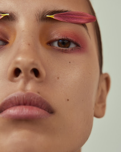 Close-up of a woman's face with petals above her eyebrows. She has brown eyes and pink eyeshadow, with a fair complexion and light pink lips.