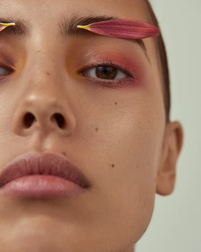 Close-up of a woman's face with petals above her eyebrows. She has brown eyes and pink eyeshadow, with a fair complexion and light pink lips.
