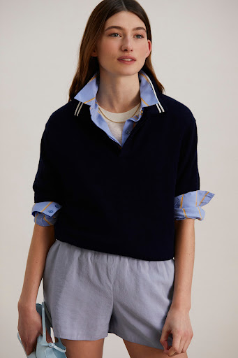 A woman in a navy short-sleeved sweater, light blue shorts, and a collared shirt holds a matching blue bag in a studio shot.