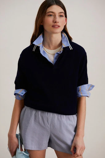 A woman in a navy short-sleeved sweater, light blue shorts, and a collared shirt holds a matching blue bag in a studio shot.