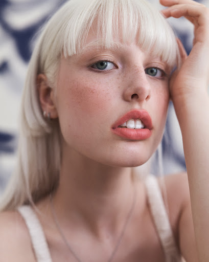 Close-up portrait of a fair-skinned adult female with blonde hair, wearing light makeup and a necklace, against a blurred background.