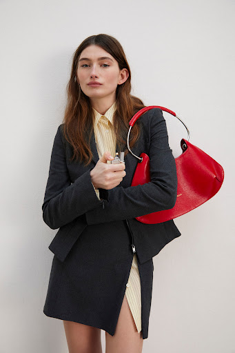 Model in dark grey suit jacket and skirt holding a bright red handbag against plain white wall