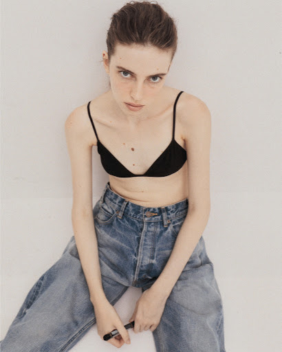 Model wearing a black bralette and blue denim jeans seated against a plain white background with make-up by Nolwenn Quintin.