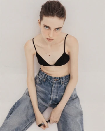 Model wearing a black bralette and blue denim jeans seated against a plain white background with make-up by Nolwenn Quintin.