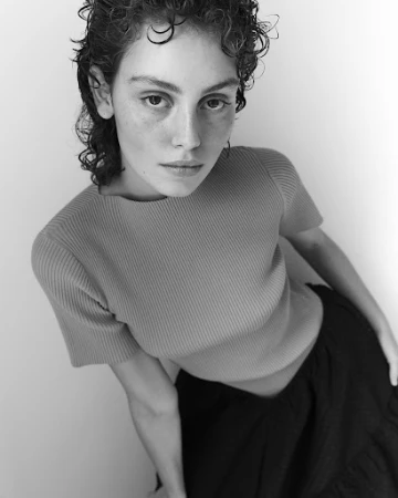 Portrait of a woman with curly hair wearing a short-sleeve ribbed top and dark skirt against a plain white background.
