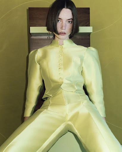 A young, fair-skinned woman with a bob haircut poses in a light yellow, tailored satin suit against a chartreuse and brown wall.