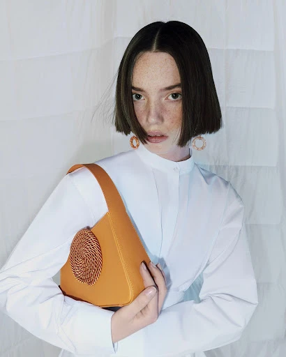A young woman with short brown hair holds a tan leather handbag while posing indoors with a neutral expression and white backdrop.