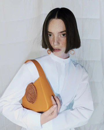 A young woman with short brown hair holds a tan leather handbag while posing indoors with a neutral expression and white backdrop.