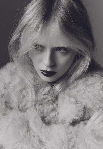 Black and white portrait of a blonde woman wearing a fuzzy coat with dark lipstick, posing in front of a grey background.