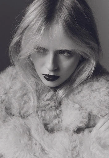 Black and white portrait of a blonde woman wearing a fuzzy coat with dark lipstick, posing in front of a grey background.