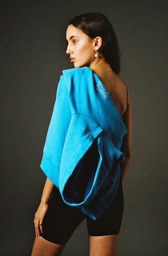 Young woman in black biker shorts and a bright blue sweatshirt draped over her shoulder poses in a studio against a grey backdrop.