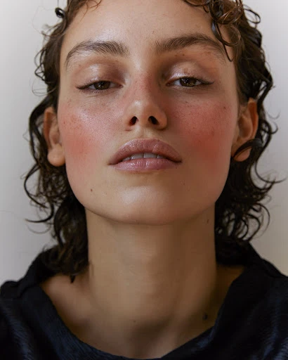 Close-up portrait of a young adult woman with wet, curly brown hair, wearing a black top, and posing against a white background.