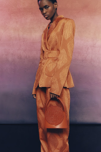 Fashionable young Black woman in a bronze-colored suit and matching structured purse poses against a mottled gradient background.