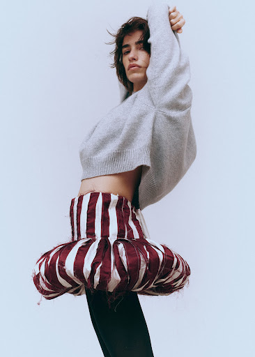 A person posing in a grey sweater and a red-and-white striped skirt in front of a light grey background in a fashion shot.