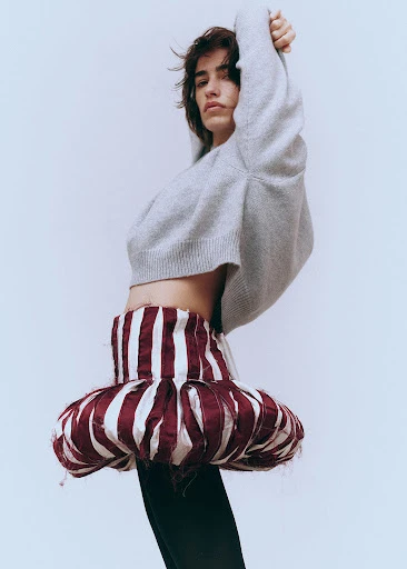 A person posing in a grey sweater and a red-and-white striped skirt in front of a light grey background in a fashion shot.