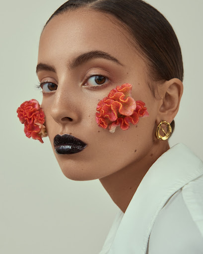 Beauty portrait of a young woman with dark lipstick and flowers on her face in a studio, wearing a white shirt and gold earrings.