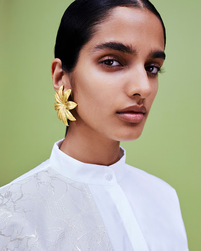 Close-up portrait of a woman with olive skin wearing a floral white shirt and gold floral earrings against a green background.