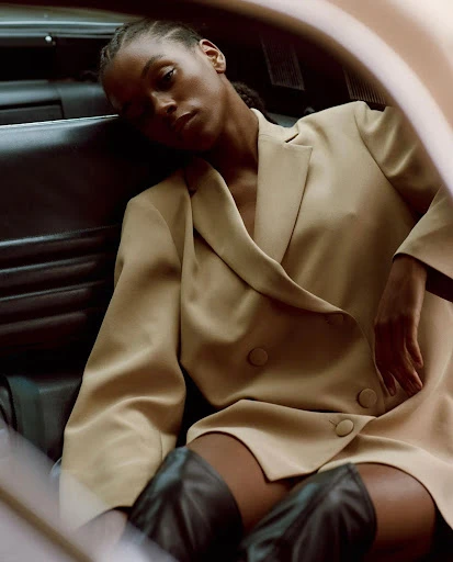 Fashion portrait of a young adult Black woman in a tan blazer dress and black boots sitting inside a car with black leather interior.