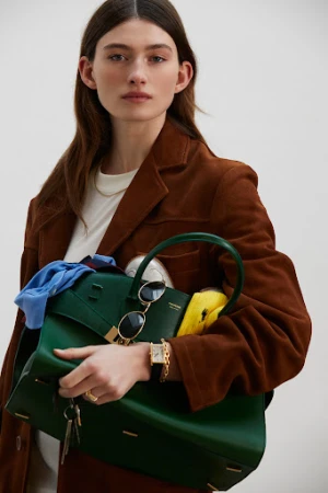 Woman in brown coat holding a large green handbag filled with items against a plain background