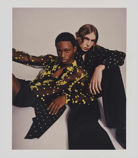 Two fashion models strike a pose, one seated and the other leaning in, wearing patterned sheer shirts and dark pants on a light background.