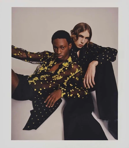 Two fashion models strike a pose, one seated and the other leaning in, wearing patterned sheer shirts and dark pants on a light background.