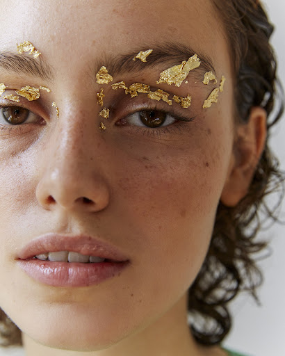 Close-up of a model's face with gold leaf applied to her eyebrows, accentuating her brown eyes and light skin in a beauty shot.