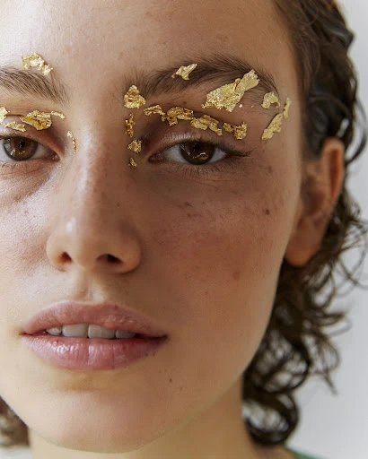 Close-up of a model's face with gold leaf applied to her eyebrows, accentuating her brown eyes and light skin in a beauty shot.