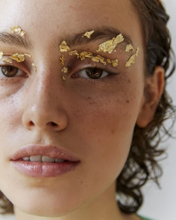 Close-up of a model's face with gold leaf applied to her eyebrows, accentuating her brown eyes and light skin in a beauty shot.