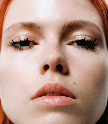 Close-up portrait of a young woman with light skin, ginger hair, and dark mascara in a brightly lit indoor setting.