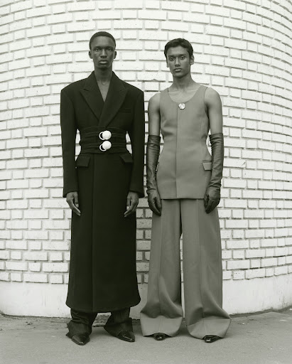 Two people stand in fashionable outfits against a textured brick wall. One wears a long black coat with a cinched waist, the other a vest top.