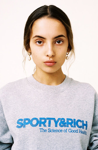 Close-up portrait of a young adult woman with light brown hair, wearing a gray sweatshirt with blue 'Sporty & Rich' text on a white background.