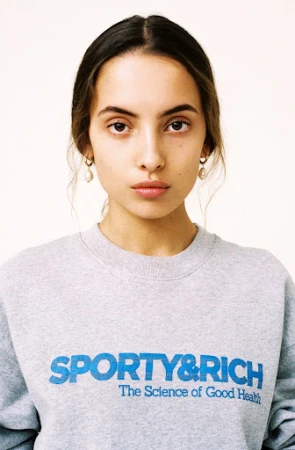 Close-up portrait of a young adult woman with light brown hair, wearing a gray sweatshirt with blue 'Sporty & Rich' text on a white background.