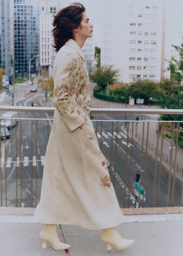 Fashionable young adult in a long, light-colored, floral-patterned coat walking on an overpass with a city street backdrop.