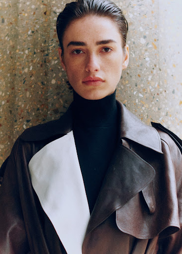 A brunette woman in a black turtleneck and stylish leather trench coat stands in front of a mottled, textured wall in a close-up shot.