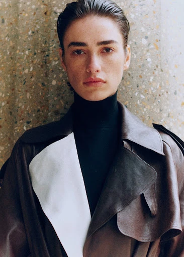 A brunette woman in a black turtleneck and stylish leather trench coat stands in front of a mottled, textured wall in a close-up shot.