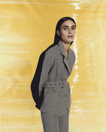 Woman in houndstooth blazer and pants posing against a yellow backdrop, creating a fashion-forward and visually striking composition.